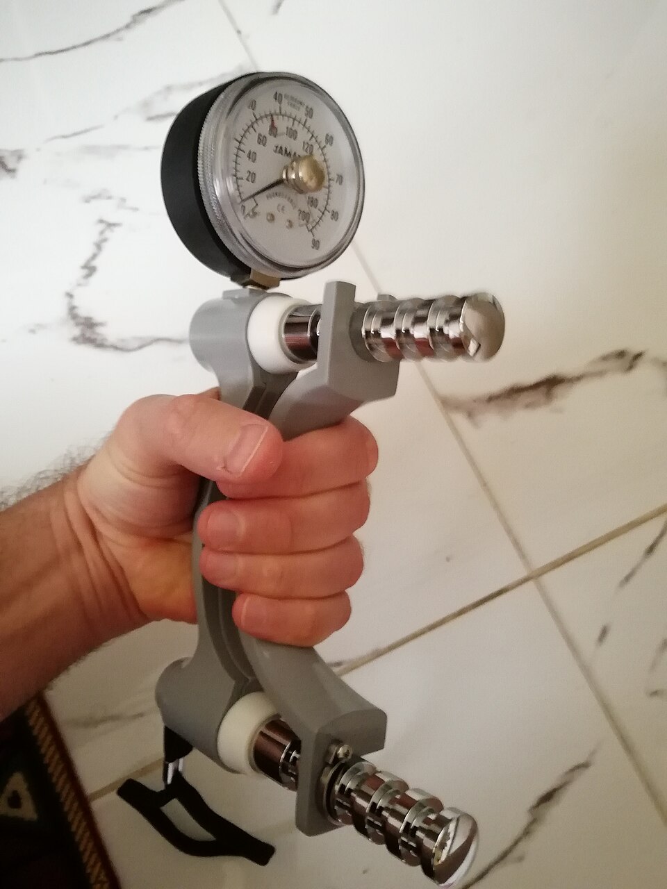 Close-up of a hand gripping a Jamar hydraulic dynamometer, with its handle and round force gauge visible.